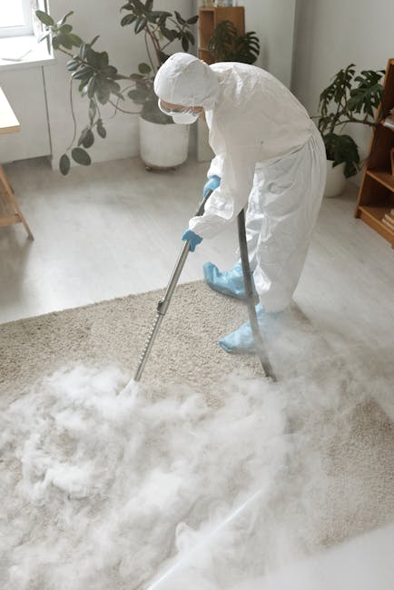 A yellow canister vacuum cleaner with a black hose and metal wand attachment is positioned on a patterned area rug in a living room. The rug lies on a wooden floor, adjacent to a black metal and wood 